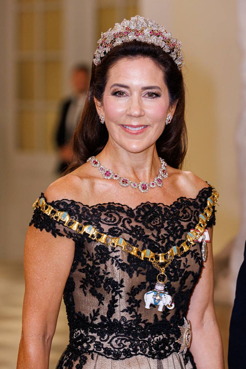 Foto colorida de mulher com joias e tiara de pedras preciosas. Ela usa uma faixa e um vestido de gala - Metrópoles