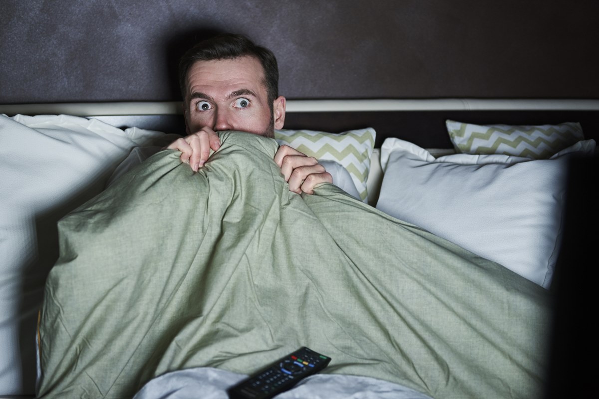 Foto de homem deitado em uma cama, com travesseiros. Ele cobre parte do rosto com um edredom e está com a cara de assustado - Metrópoles