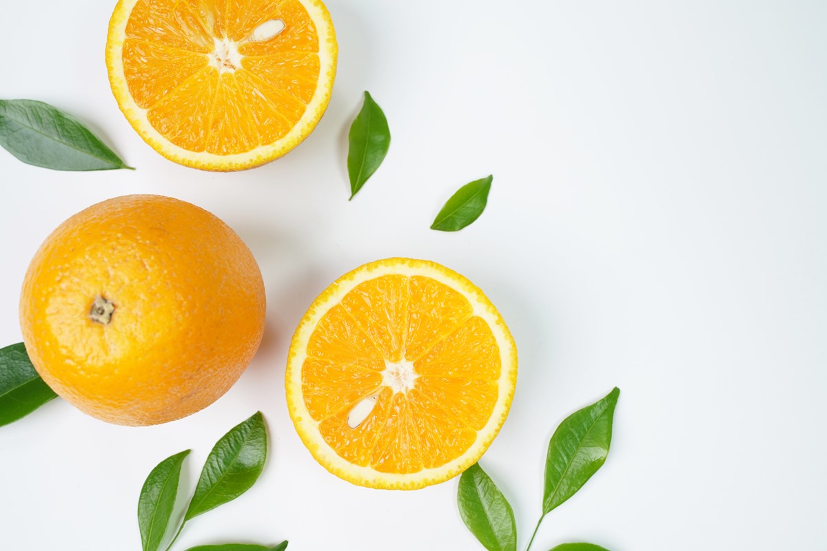 Foto de uma laranja cortada ao meio e de uma fruta inteira sobre uma superfície branca. Ao redor tem folhas de laranjeira - Metrópoles