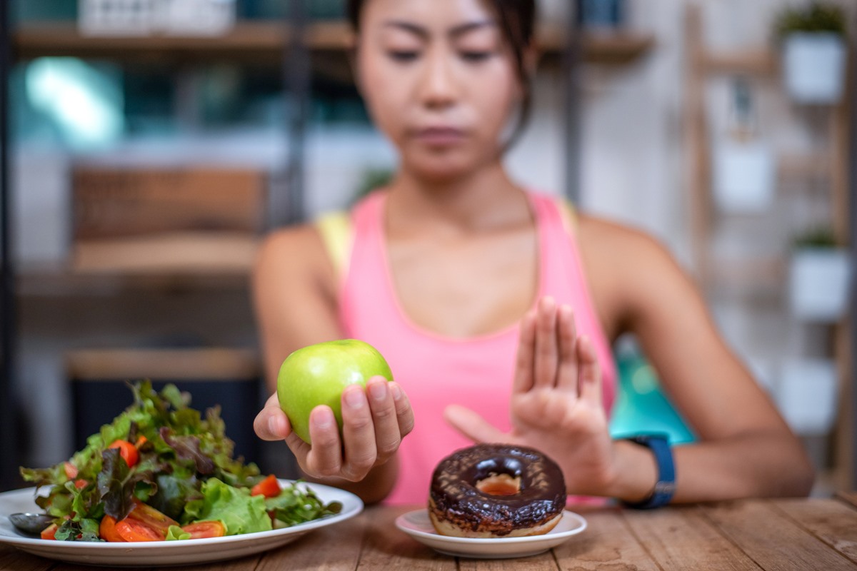 Foto de mulher com maçã verde na mão. Ela está escondendo com a outra mão um prato com um donut. Do lado dela tem um prato com salada de alface - Metrópoles - alimentação - hábitos para evitar câncer