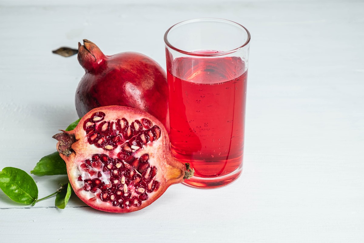 Foto colorida de romã inteira e um cortada pela metade ao lado de copo transparente de suco - Metrópoles