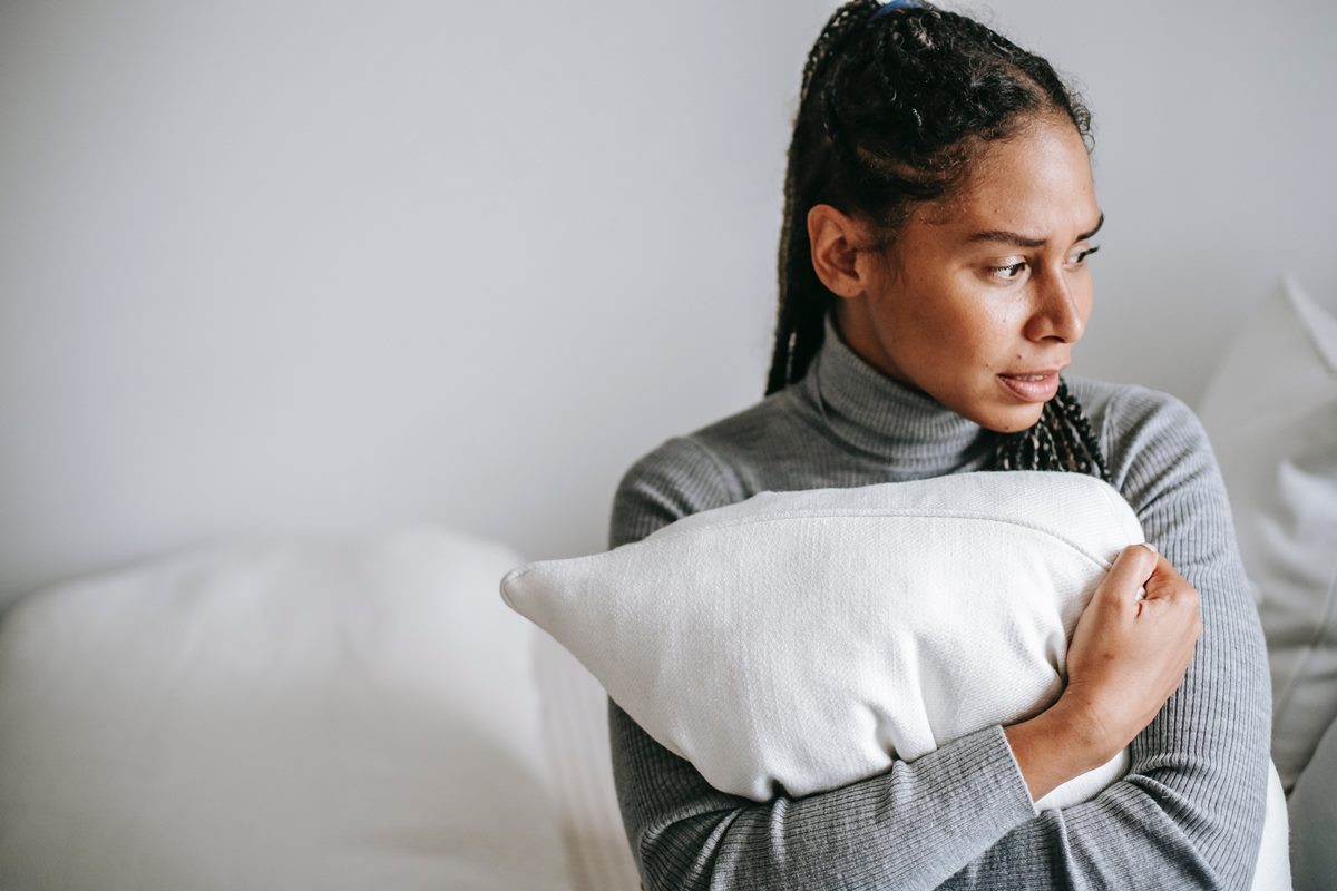 Mulher negra vestida com blusa cinza abraçada em um travesseiro com cara de preocupação