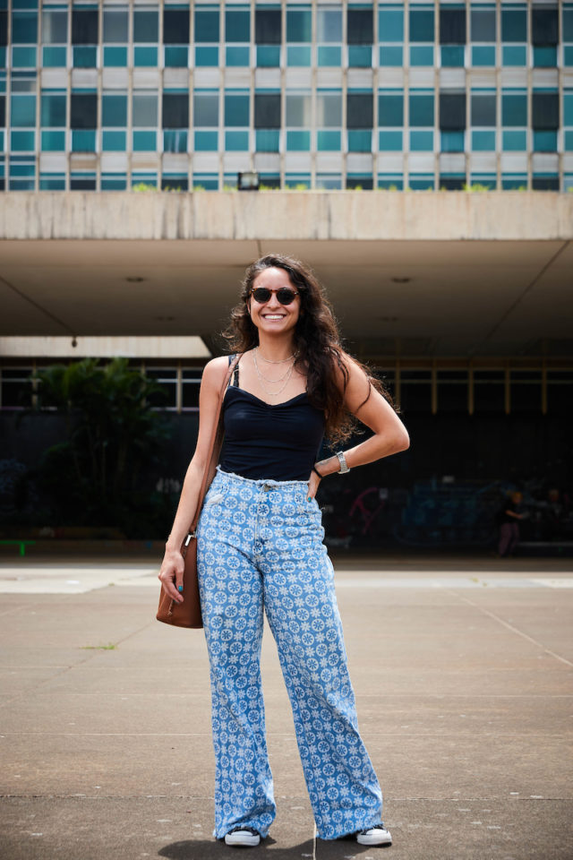 Mulher com sorriso posa para a foto. De regata preta, ela carrega sua bolsa marrom ao lado direito encostada em sua calça jeans azul estampada com flores brancas junto a um tênis preto com detalhes brancos.