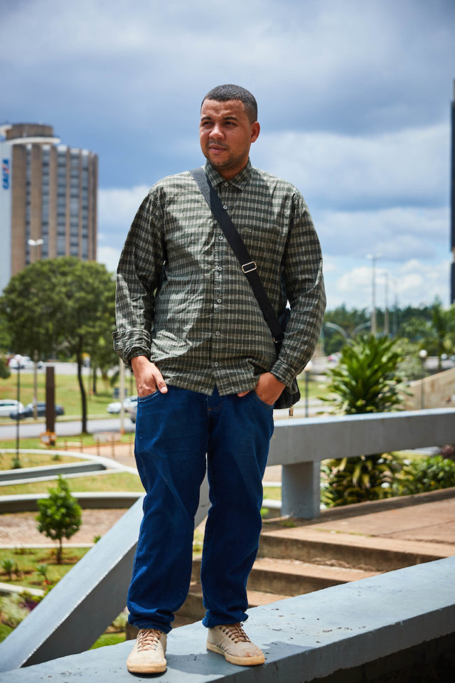 Homem negro posa para a foto. Com uma camisa de botão xadrez em verde, ele carrega uma bolsa tranversal. Suas mãos ficam postas dentro do bolso de sua calça jeans azul que encostam em seus tênis em tons terrosos.