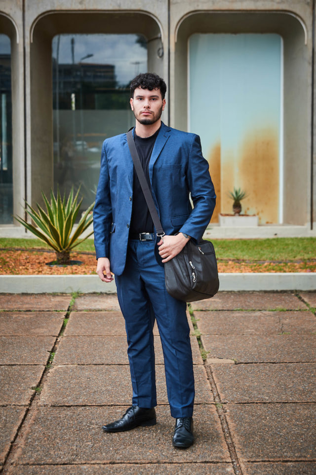 Homem em terno azul marinho com camisa preta e bolsa transversal na cor marrom, fotografado em frente a galeria.