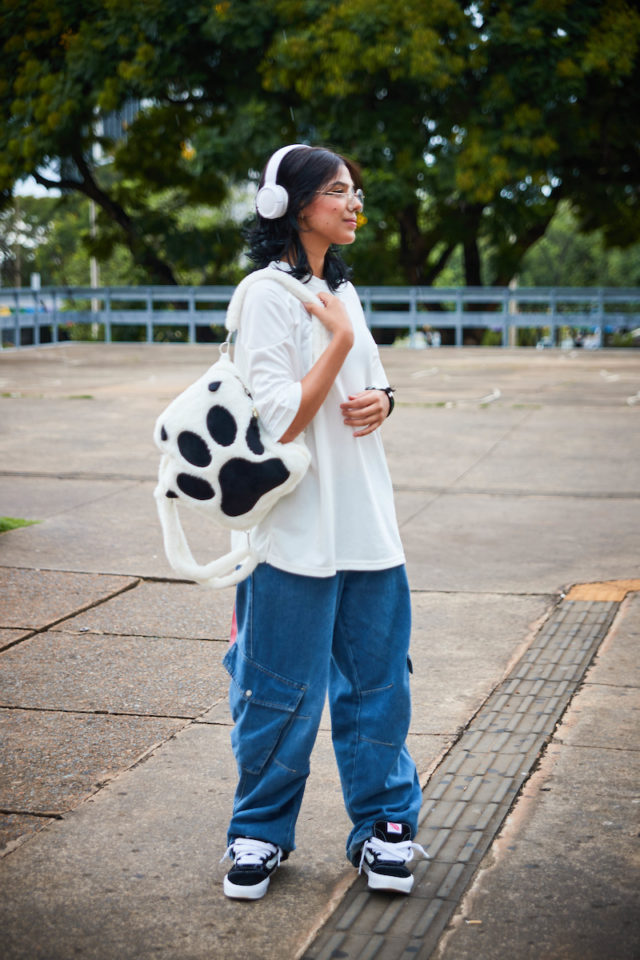 Garota com fones brancos posa para a foto. Carregando uma mochila com formato de pata em seu ombro direito, a jovem veste uma camiseta branca lisa, junto a uma calça jeans larga e calçado preto com detalhes em branco.