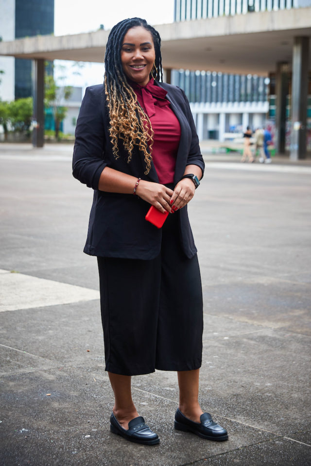 Mulher negra posa para a foto. Com cabelo em locs, ela usa um conjunto de blazer e calça em preto, junto a uma blusa vinho