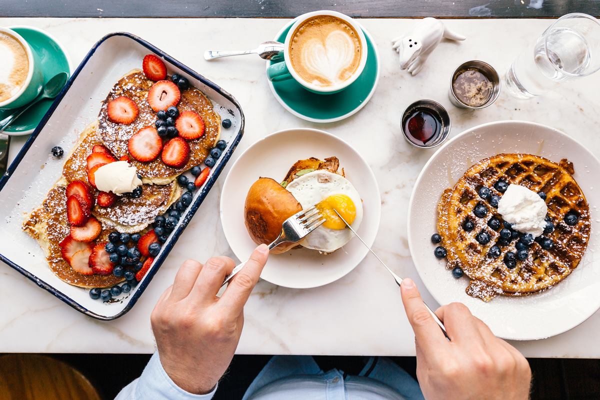 Foto de mãos masculinas cortando um ovo dentro de um sanduíche. Ao lado, tem tigelas com waffles e xícaras de café e capuccino - Metrópoles