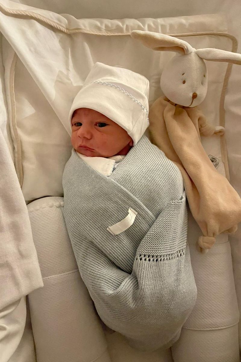 Foto de bebê com touca e roupa branca. Ele está enrolado em uma manta azul e ao lado de um coelho de naninha - Metrópoles