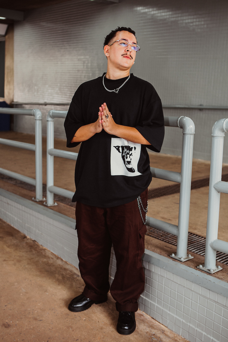 De mãos postas e olhando para fora, o homem posa para a foto. De camiseta preta e calça marrom escura, o rapaz combina a camisa com seus sapatos também em preto.