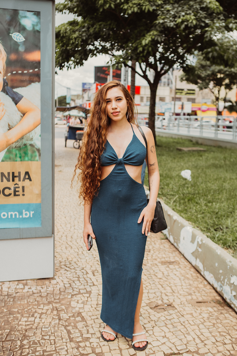 Mulher de cabelo ruivo em vestido verde. Com sua bolsa preta no ombro esquerdo e celular na mão direita, a jovem posa para a foto.