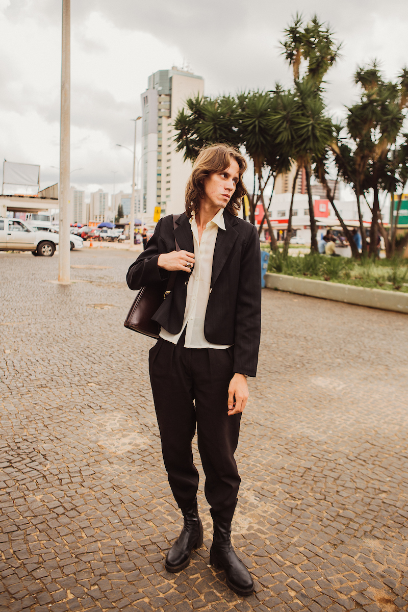 Homem com conjunto de alfaiataria posa para foto em frente a praça. Com sua bolas apoiada em seu ombro direito, o rapaz combina o conjunto em preto com seu coturno de mesma cor.