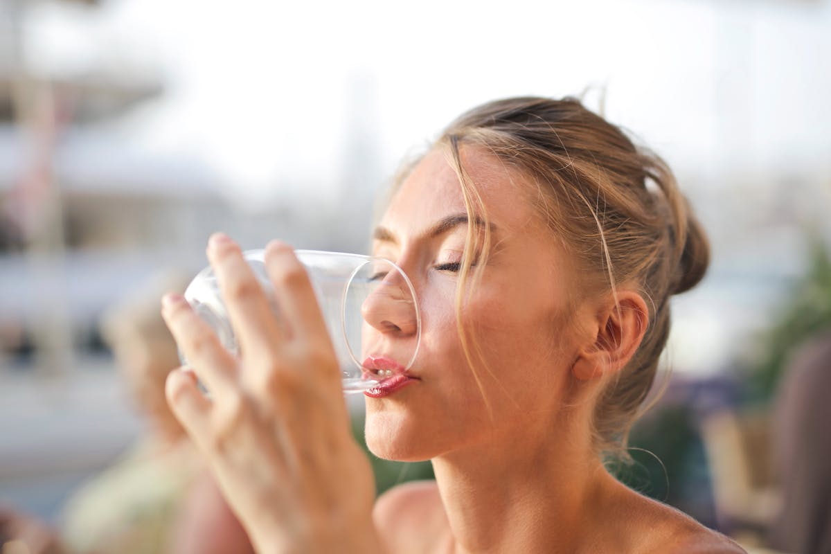 Jovem mulher loira bebendo água em copo de vidro transparente - Metrópoles