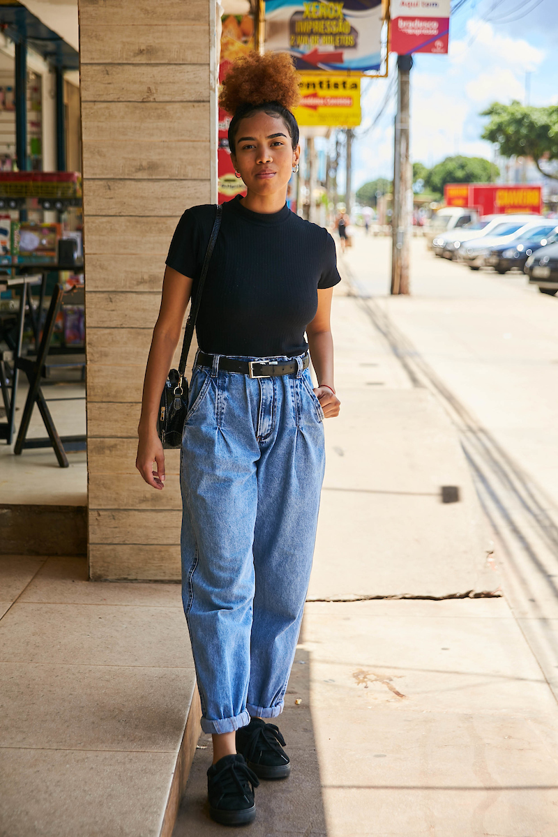 Jovem de camisa preta posa para foto. Com sua bolsa preta apoiada no ombro direito, ela veste uma calça jeans segurada por um cinto também preto. 