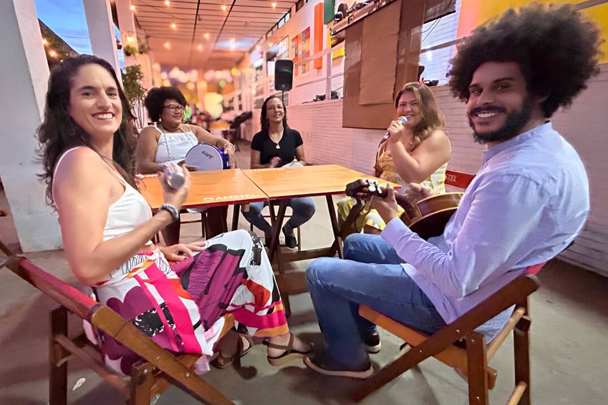 foto colorida de grupo de samba formado por quatro mulheres e um homem sentados ao redor de mesa de madeira - metrópoles