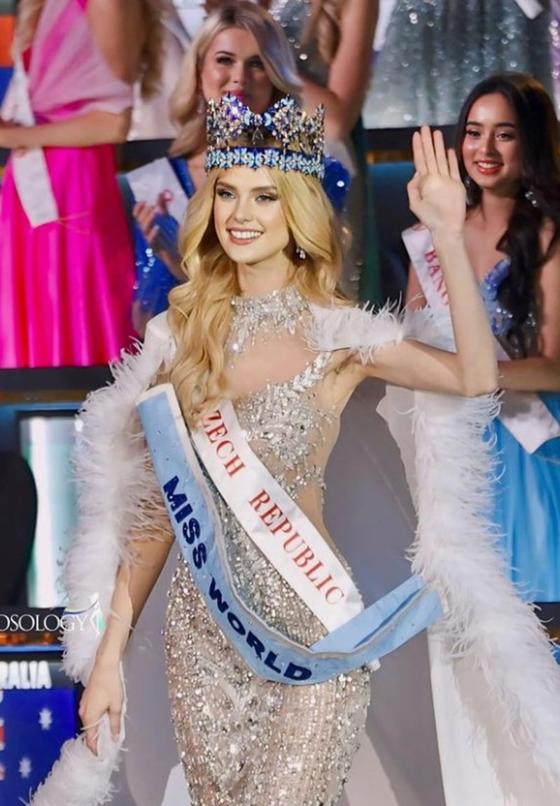 Na imagem colorida, uma mulher usando vestido brilhoso acena enquanto carrega a faixa e a coroa de Miss Mundo 2024 - Metrópoles