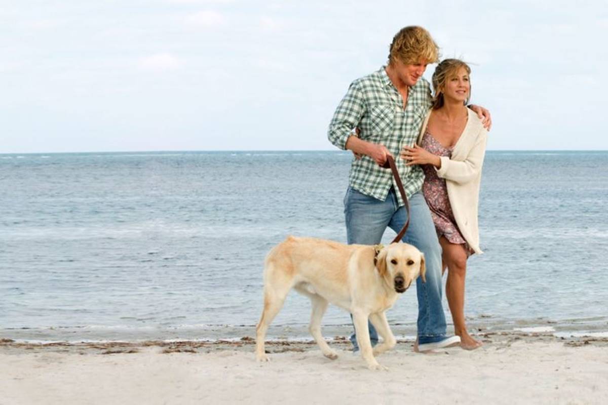 Foto colorida de Marley e Eu. Imagem mostra duas pessoas na praia, andando com um cachorro na coleira - Metrópoles