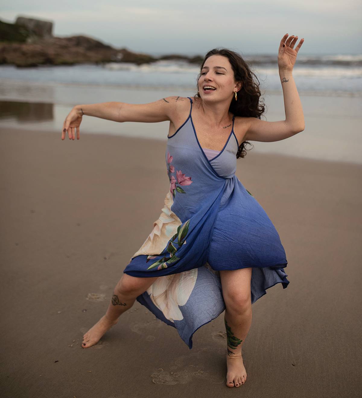 Foto colorida de uma mulher com o vestido estampado dançando descalça na areia em frente ao mar - Metrópoles