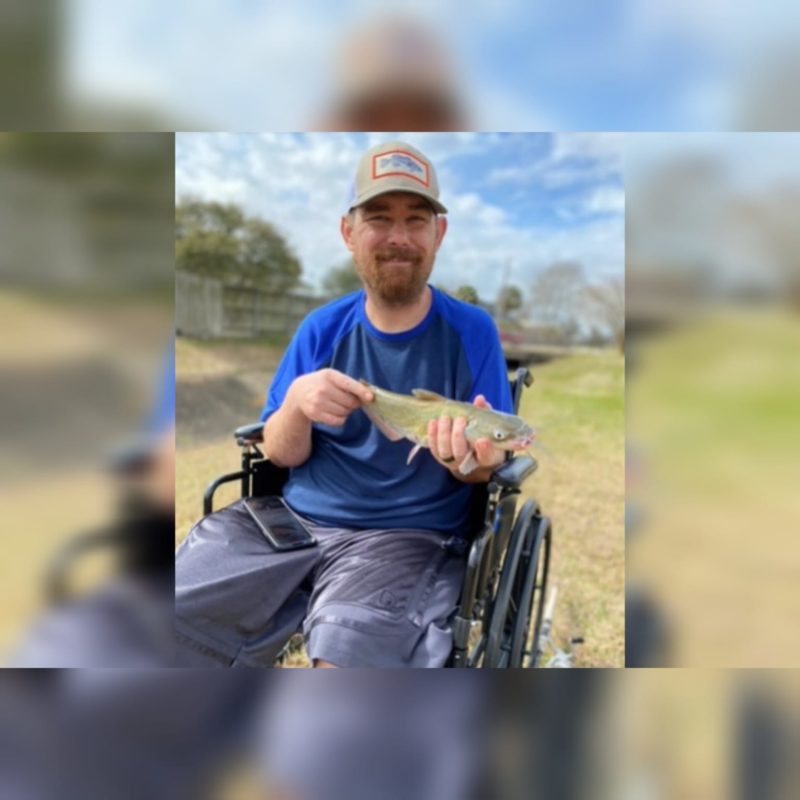 Um homem branco de camisa azul sentando em uma cadeira de rodas segurando um peixe