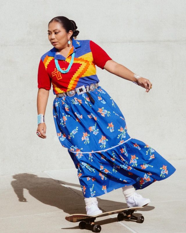 Naiomi é fotografada andando de skate. Na imagem, ela aparece vestindo uma saia azul com flores estampadas. Sua camisa traz as cores vermelho, amarelo, azul e laranja junto ao colar, brinco e pulseias na cor azul.