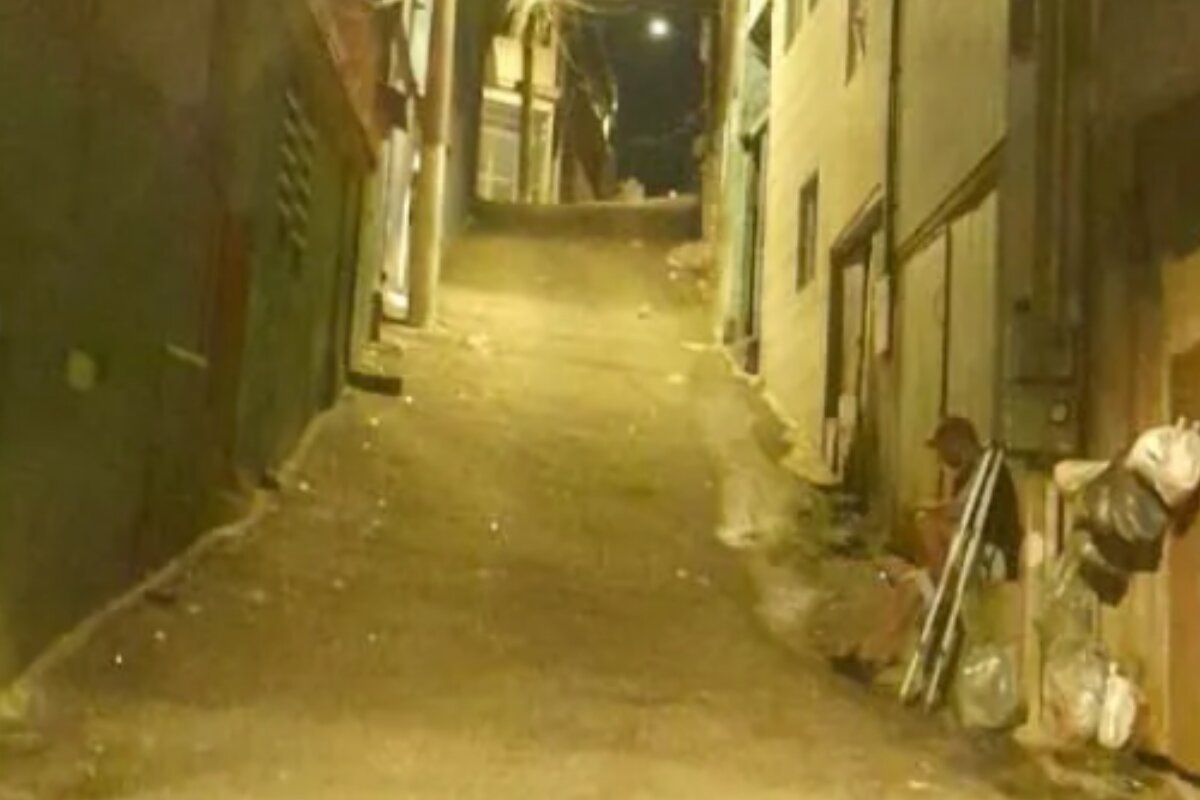 Foto colorida de homem sentando em frente de residência humilde, em rua de terra, do lado do qual há um par de muletas - Metrópoles