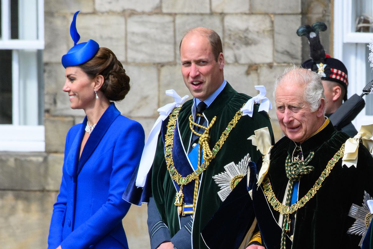 Foto de mulher com roupa azul ao lado de dois homens com trajes da realeza britânicos - Metrópoles