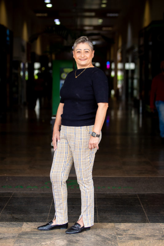 Mulher posa para foto. Com sua camiseta preta, ela sorri para imagem vestindo uma calça xadrez nas cores preto, amarelo e branco, combinando com seu sapato mocassim preto.