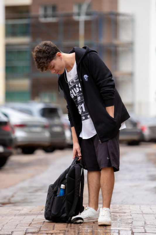 Rapaz posa para foto. Olhando para baixo enquanto puxa sua mochila preta, ele põe sua mão esquerda no bolso do casaco de zíper aberto, que deixa espaço para sua camisa branca com estampa em preto. Na parte inferior ele usa uma bermuda preta e um tênis branco.