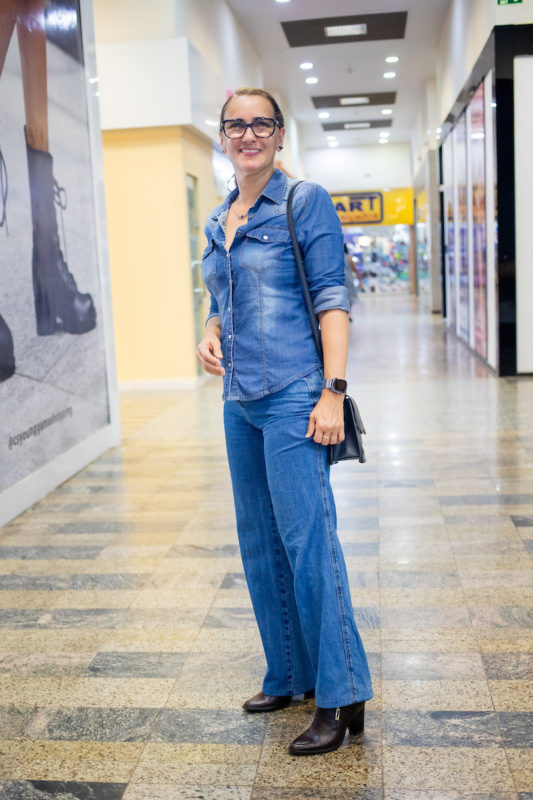 Mulher posa para foto. Vestindo um conjunto jeans, combinando uma bolsa preta e bota marrom, ela sorri para imagem em centro comercial.