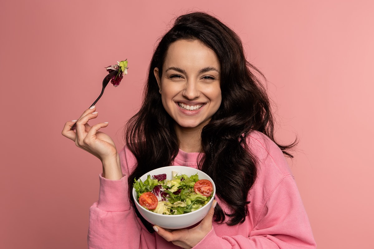 Foto colorida de mulher jovem segurando um garfo e um pote com salada dentro - Metrópoles