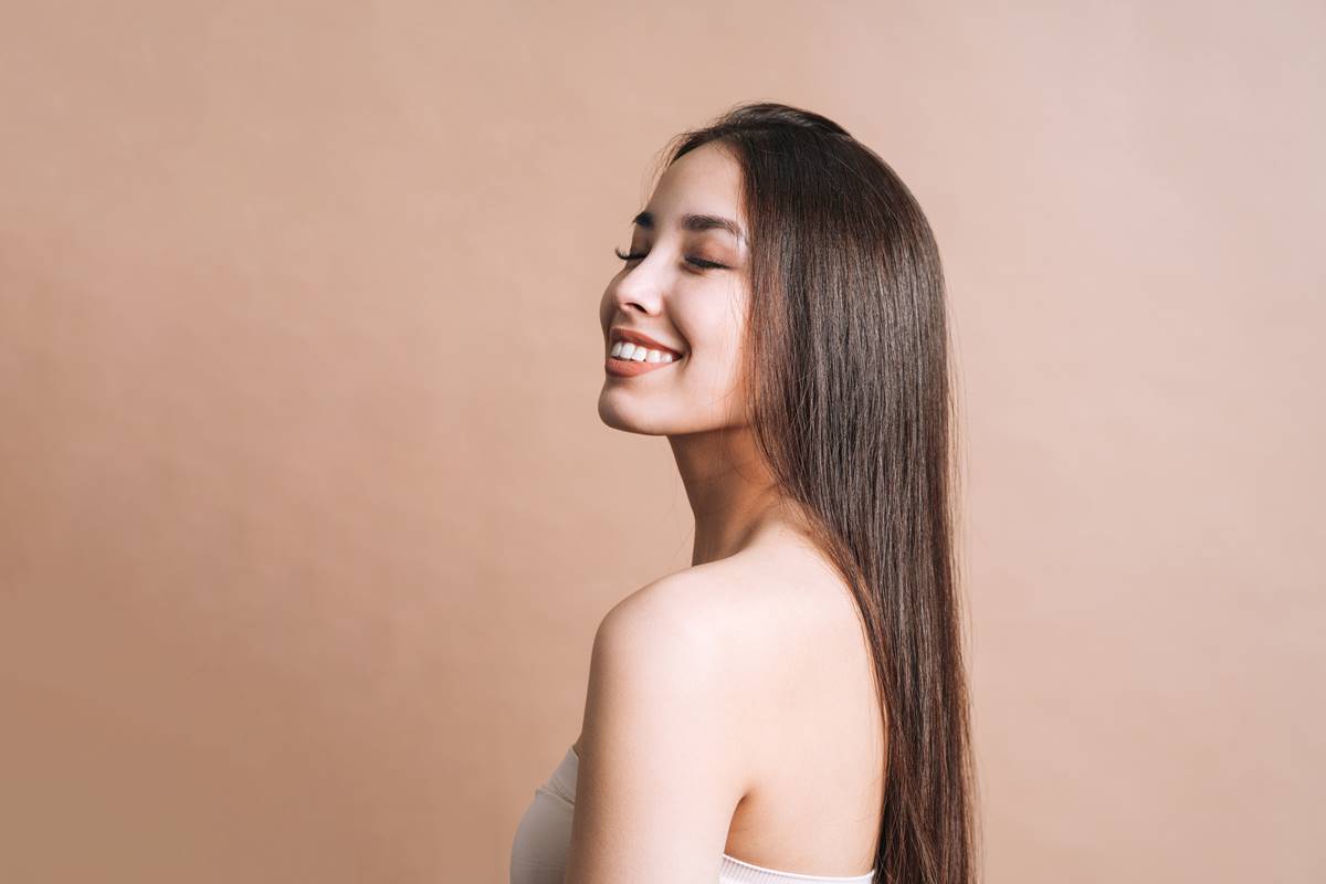 Foto colorida de uma mulher branca com o cabelo liso e castanho sorrindo em um fundo bege - Metrópoles