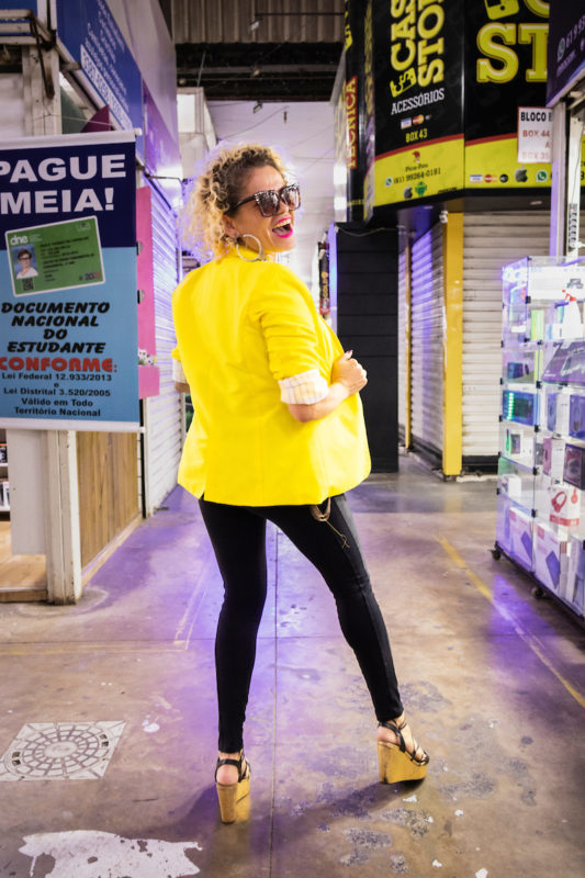 Mulher posa para a foto. Com seu blazer amarelo, ela aposta em um look de camiseta e calça preta, junto á um salto. Nos acessórios, ela usa um brinco e óculos escuro.