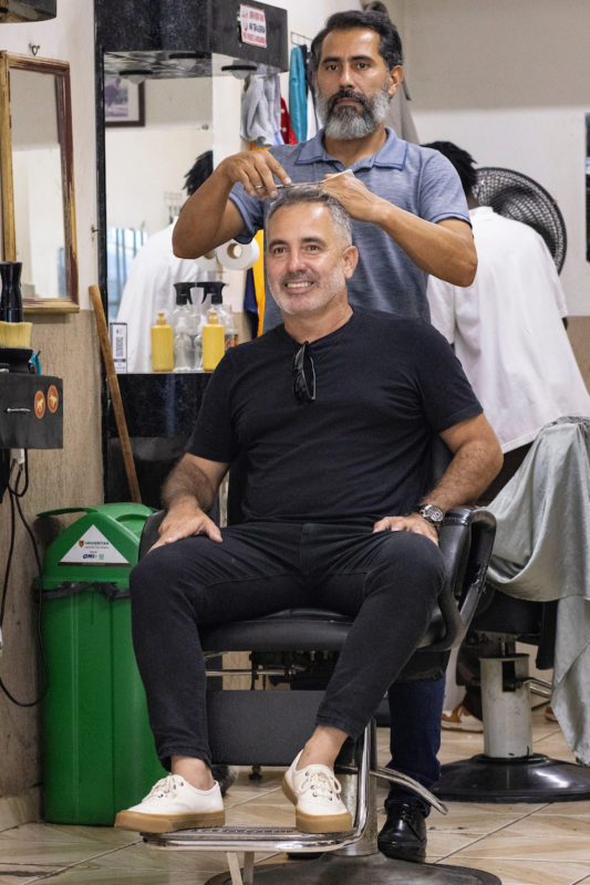 Homem sorri enquanto corta o cabelo. Em um visual justo ao corpo, ele investe no preto na camiseta e calça e deixa o destaque para o calçaco branco com a sola marrom.