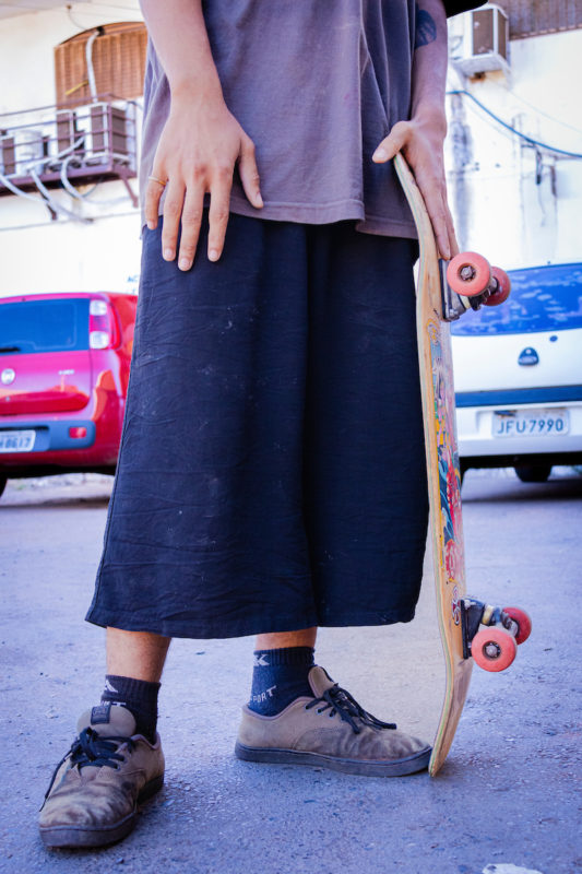 Rapaza posa para foto que mostra sua calça acima do tronozelo. Apoiando seu skate com a mão, o jovem calça um tênis preto de camurça junto a uma meia de cano médio.