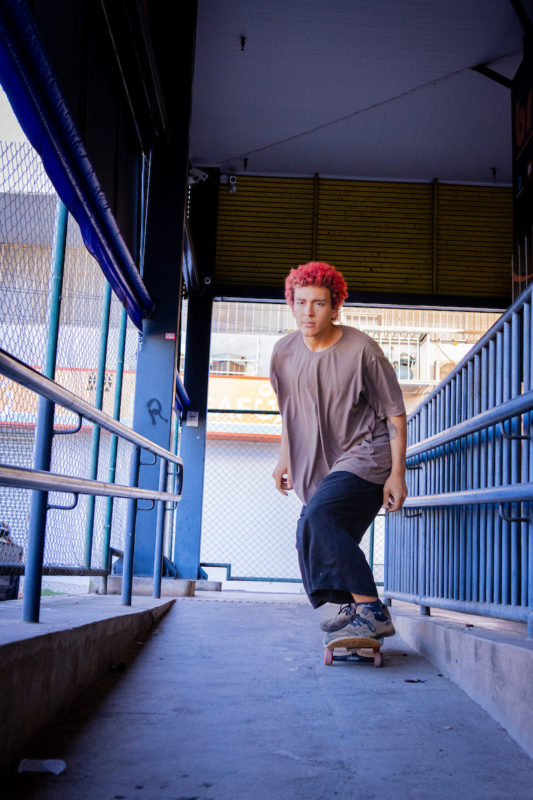 Rapaz anda de skate com uma camisa marrom larga e calça preta acima do tornozelo.