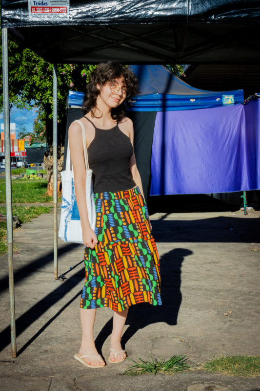 Garota posa para foto. Com sua camiseta regata preta e saia rodada colorida nas cores vemelho, azul, verde e amarelo ela sorri com sua bolça branca com estampa azul.