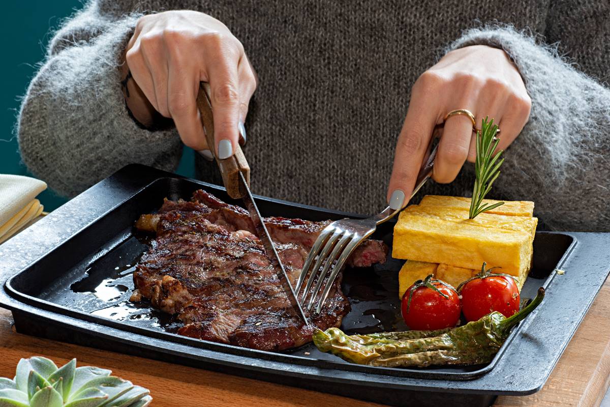 Foto colorida de mão de mulher cortando carne - Metrópoles