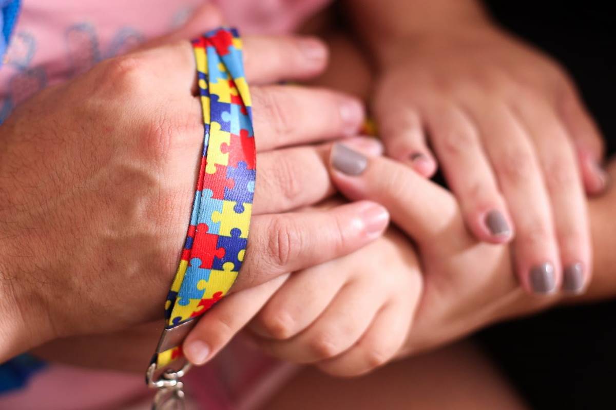 Foto colorida de pais segurando a mão de criança com o cordão de autismo