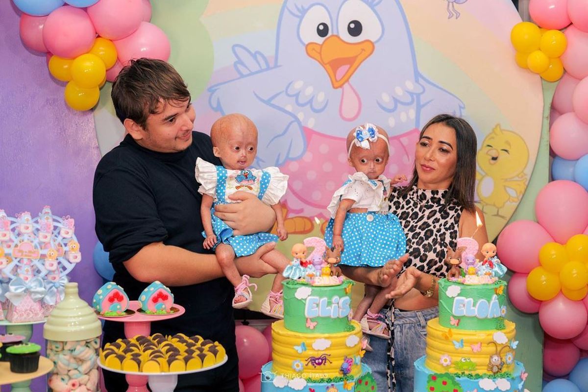 Foto colorida de homem e mulher, cada um segurando uma bebê. Eles estão em uma festa infantil com o tema Galinha Pintadinha - Metrópoles