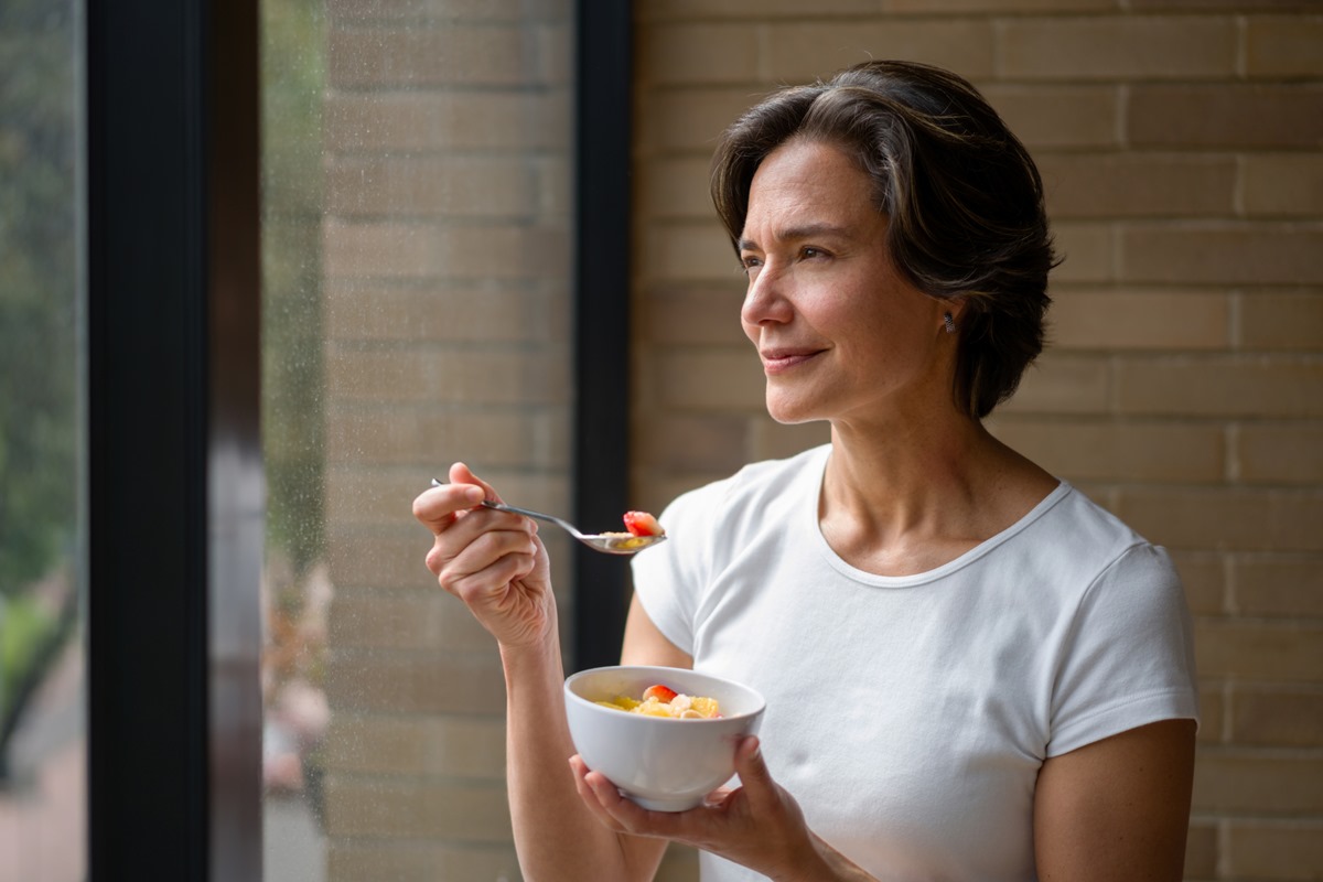 Foto colorida de mulher olhando pela janela enquanto come morangos - Metrópoles