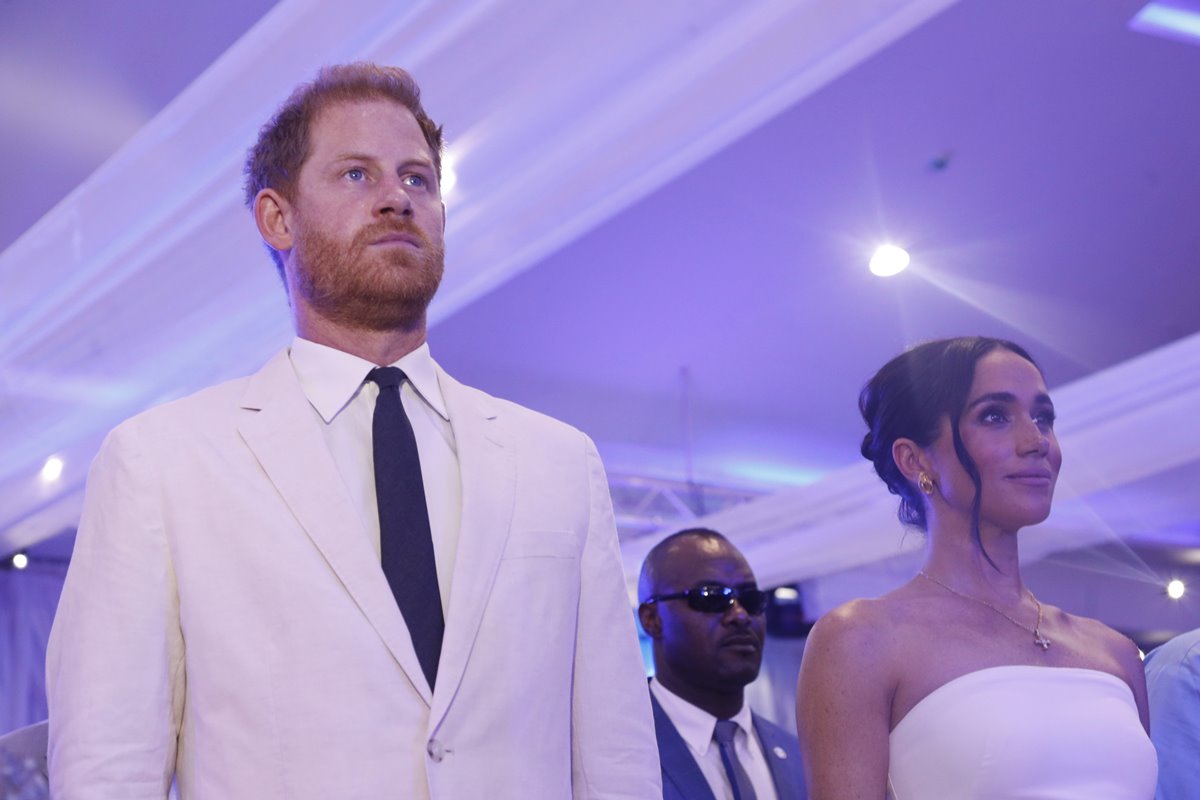 Foto colorida de homem ruivo com blazer branco e rosto sério. Ele está ao lado de mulher com cabelo preso e roupa branca sem mangas - Metrópoles