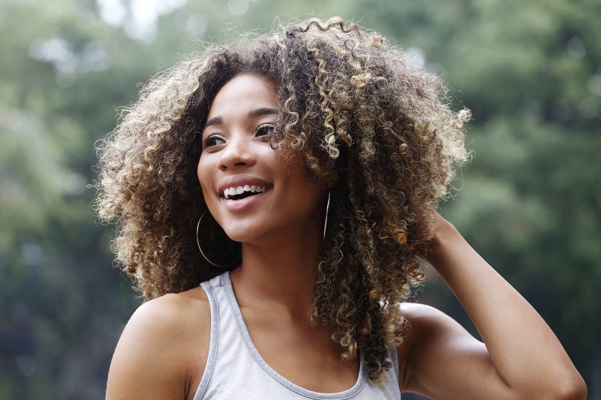 Foto colorida de uma mulher de cabeçlo cacheado sorrindo e olhando para o lado com a mão na cabeça - Metrópoles