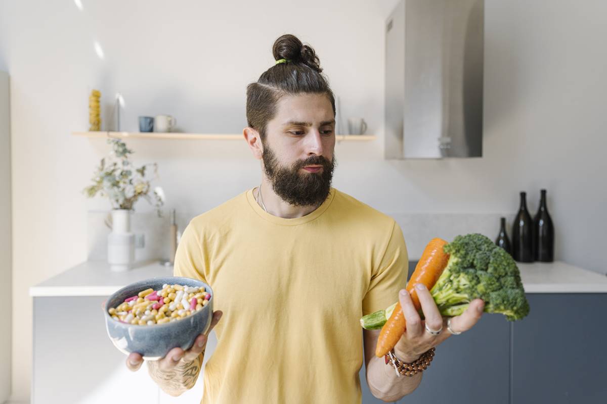 Foto colorida- Homem segurando um pote com suplementos em uma mão e, na outra, brócolis e cenoura