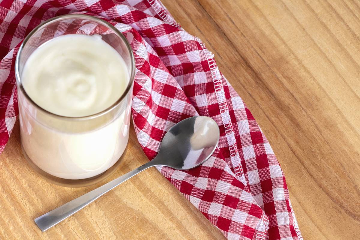 Foto colorida - Copo com iogurte, colher e pano xadrez na mesa