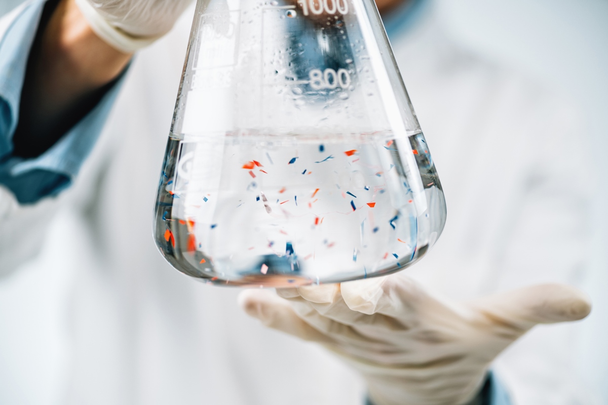 Foto colorida de vidraria de laboratorio Erlenmeyer com uma solução transparente e microplasticos. Metrópoles