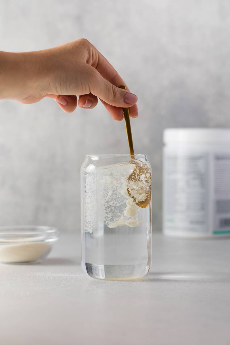 Foto colorida - Mão mexendo pó branco dentro de um copo transparente com líquido branco com colher dourada