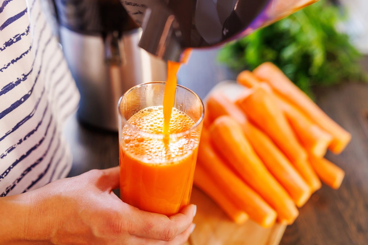 Foto mostra as mãos de uma pessoa segundo um copo com suco de cenoura. Ao fundo várias cenouras descascadas - Metrópoles