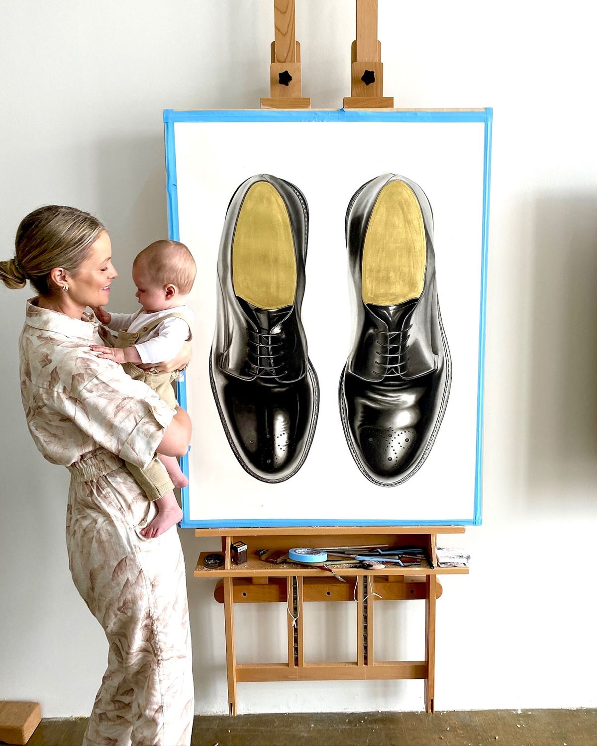 Foto colorida de mulher segurando bebê ao lado de quadro com desenho de par de sapatos - Metrópoles