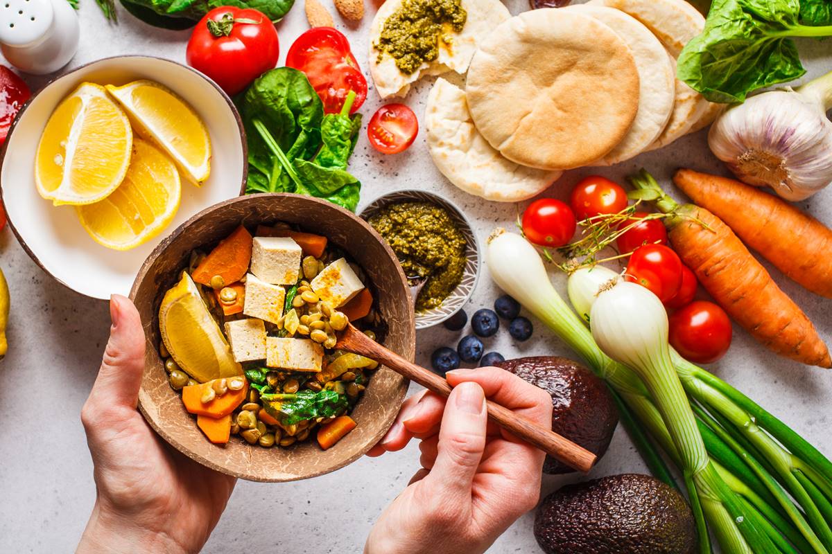 Foto colorida de mão segurando colher que está dentro de bowl com alimentos. Ao fundo, há uma mesa com vegetais, frutas e pães - Metrópoles