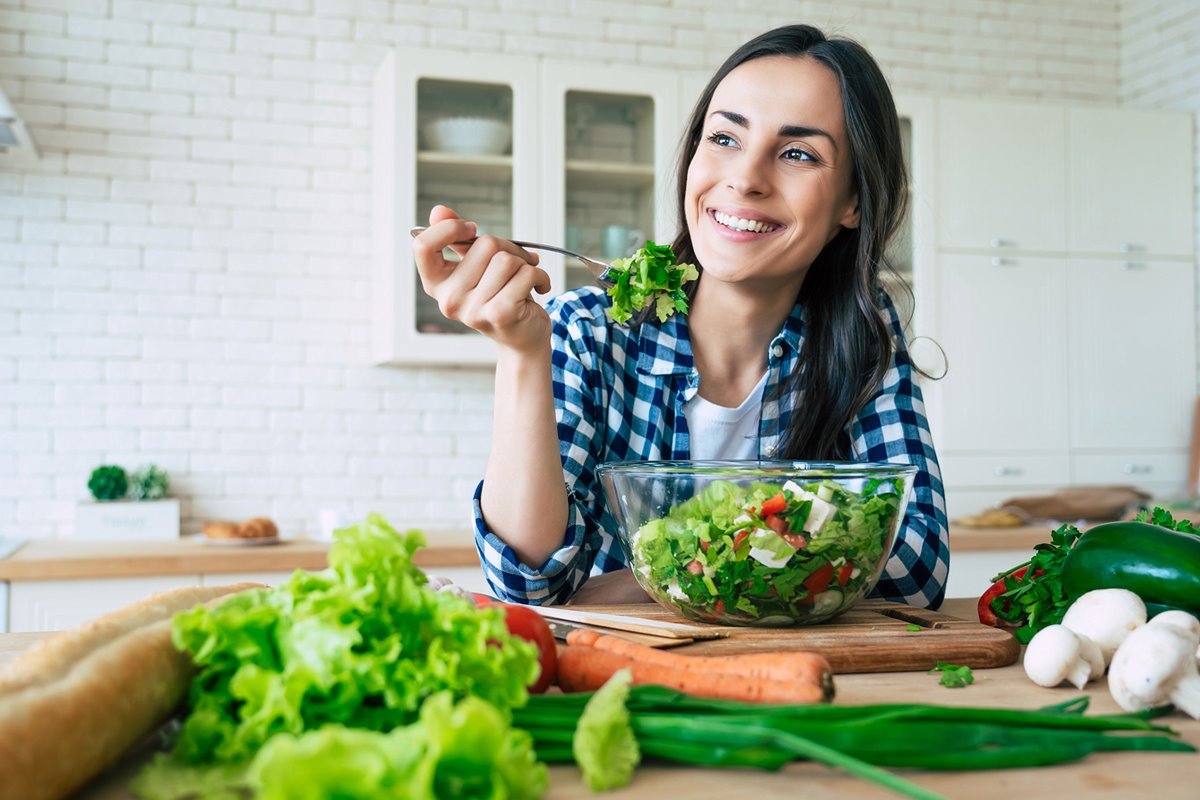 Foto colorida de mulher com cabelo castanho. Ela está comendo salada - Metrópoles